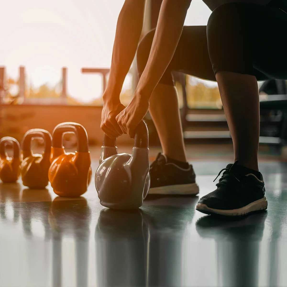 Osoba w stroju sportowym podnosząca kettlebell z podłogi na siłowni, w tle rząd kolorowych odważników w ciepłym świetle
