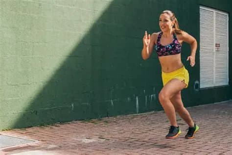Kobieta w stroju sportowym biegnie chodnikiem wzdłuż ciemnozielonej ściany w słoneczny dzień.